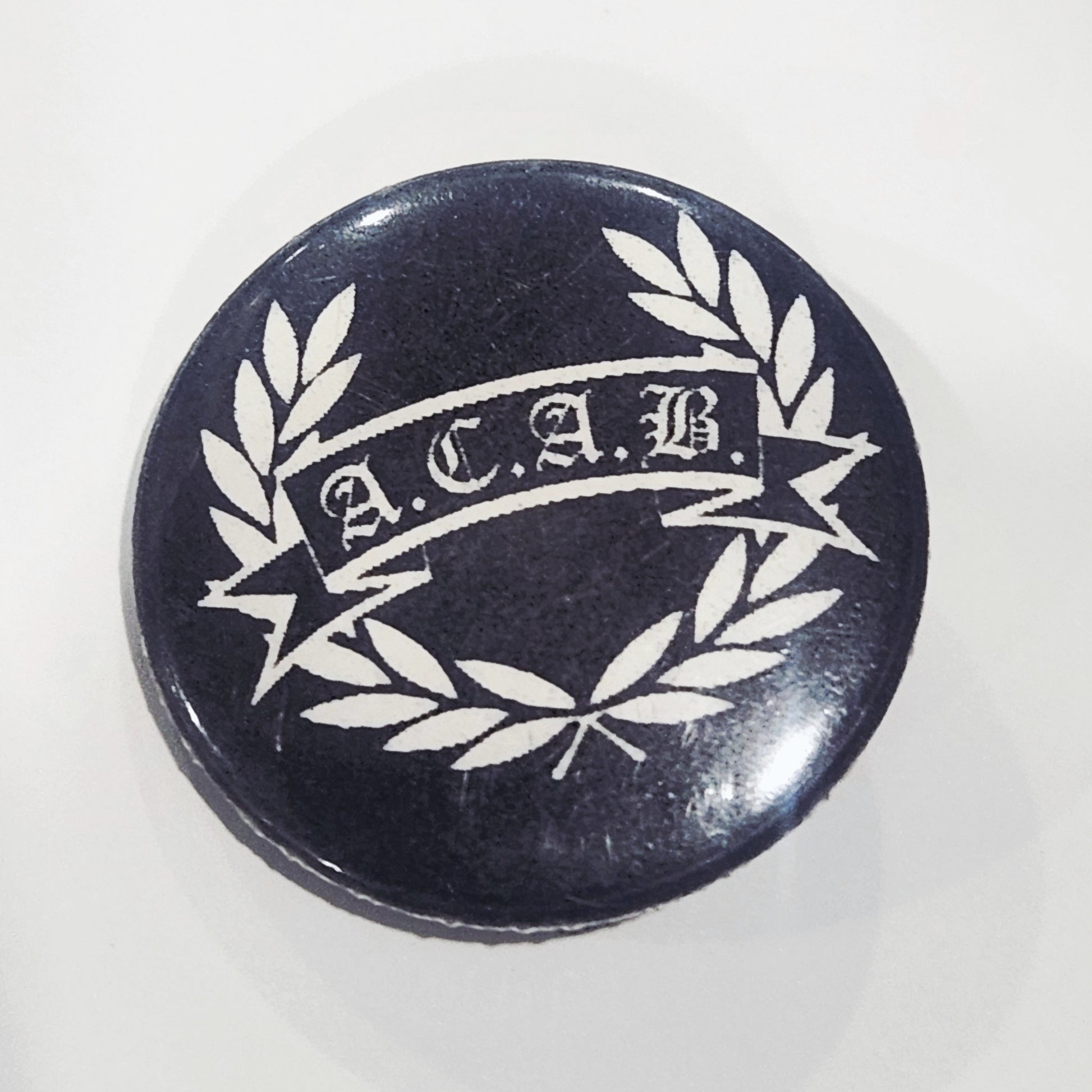 Round button badge with 'A.C.A.B.' text and laurel wreath on a black background