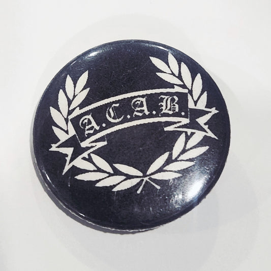 Round button badge with 'A.C.A.B.' text and laurel wreath on a black background